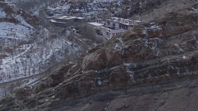 Hemis Monastery's white multi-story buildings sit among snow-dusted Himalayan slopes in Ladakh. Slow forward tracking aerial shot reveals the temple complex and surrounding valley settlements.