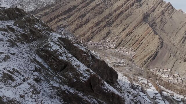 Ladakh's Hemis Monastery emerges in a valley between layered mountain ridges dusted with winter snow. The camera moves forward past rocky slopes to reveal the white Buddhist complex and remote village
