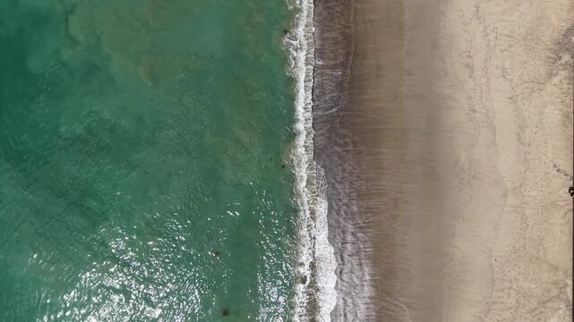 Cinematic 4K 10-bit aerial drone footage captured above Malibu Beach on a sunny day, featuring a bird&rsquo;s-eye view of ocean waves crashing onto the sandy shoreline. The drone remains steady in a hover.
