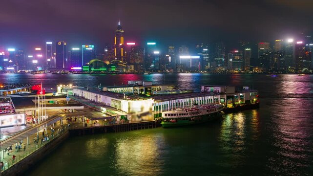 Time Lapse night view of Star Ferry Pier in Hong Kong with Victoria Harbour skyline, illuminated skyscrapers, colorful city lights reflections on water and busy urban waterfront scene
