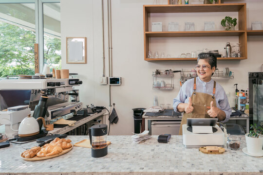 Portrait of a confident senior Asian woman, a proud small busin ess owner, giving a double thumbs up in her modern coffee shop. Concept of entrepreneurship, success, and customer satisfaction.
