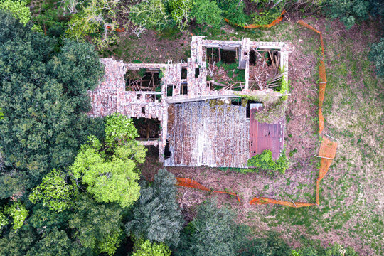 Aerial view of a ruined stone building with a collapsed roof surrounded by lush green forest and orange safety netting in Reggello, Tuscany, Italy.