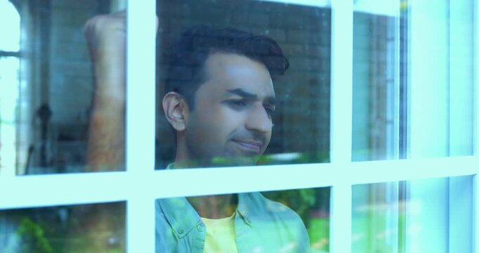 Frustrated young Indian man banging glass window while expressing intense anger and distress, viewed from outside showing reflections on the glass pane in a modern Indian home setting
