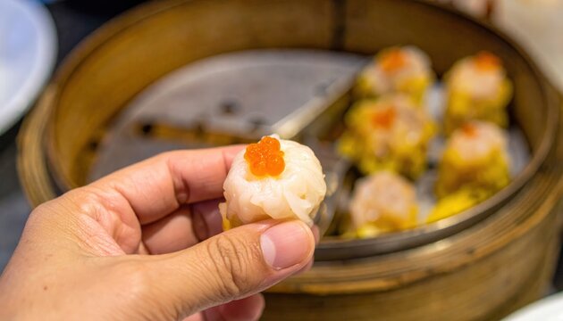 Hand holding a delicate siu mai, adorned with crab roe, ready to be enjoyed in a traditional bamboo steamer.