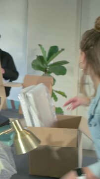 Vertical video: Approaching desk, woman in denim lifting bubblewrap chair panel at office for setup