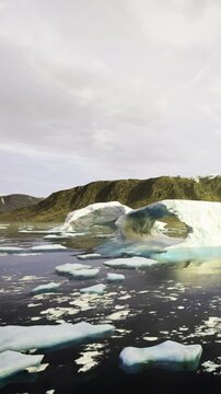 Large ice formations float in the water while rocky land rises in the background. Clouds cover the sky in this Arctic scene filled with ice and water.
