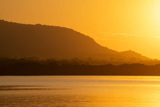 Hazy golden hour landscape featuring a tropical coastline with dense trees and distant hills.