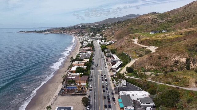 Cinematic 4K 10-bit aerial drone footage captured above the Pacific Coast Highway in Malibu, featuring a smooth reverse dolly movement over vehicles traveling along the scenic coastal highway.