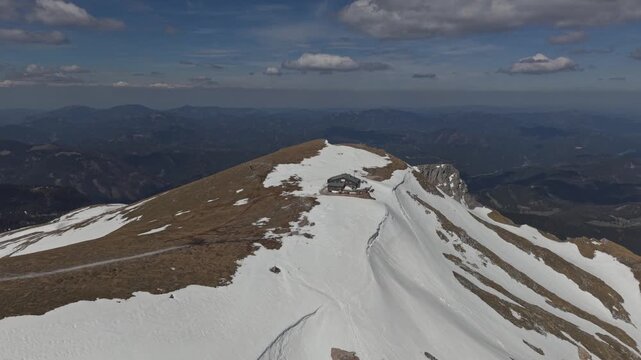 Aerial perspective of a snow-covered mountain ridge leading to a small alpine hut near the summit.