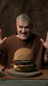 Happy excited prehistoric man having a tasty fast food meal in his cave: he grabs a big hamburger and smiles at the camera
