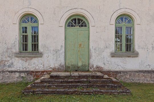 Facade of an old, weathered building featuring a central arched green metal door and two matching arched windows on either side, Muhu, Estonia, Europe.
