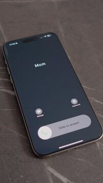 Vertical Screen: A smartphone sits on a table. An incoming call appears with Mom's name on the screen. The time is shown at the top of the phone. A simple moment at home.