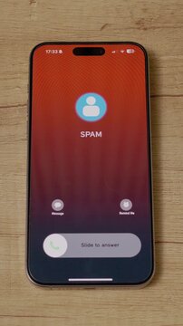 Vertical Screen: A smartphone receives an incoming call marked as spam while resting on a wooden table in a home setting. The call alert flashes on the screen, warning the user.