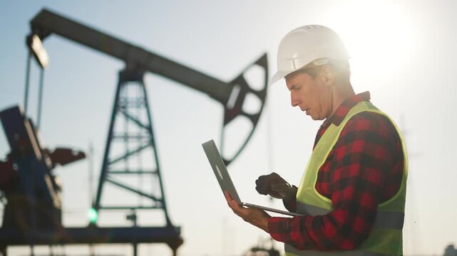 Worker checks laptop near pumpjack in oilfield engineer wearing helmet reflective vest and glove studies data while pumpjack operates at dusk conveying energy output oil industry site and safety focus