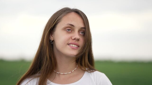 Woman closes eye in field, girl with long hair wearing shell necklace and white tshirt, gentle smile and calm expression, outdoor portrait with soft green blur behind, relaxed pose, eye closed