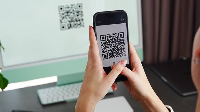 Person scanning QR code from computer screen with smartphone at home workspace. Digital interaction between devices and online service access concept.