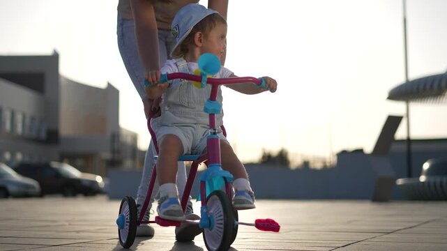 Helping toddler learn to ride tricycle with parent and mother guiding push and balance outdoor near sunset on paved plaza child wearing cap looks forward while parent supports and encourages skill