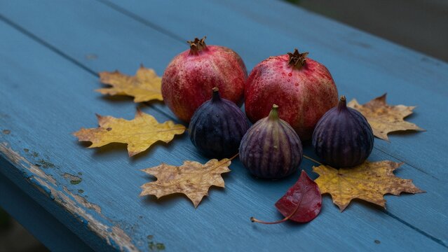 Ripe pomegranates and purple figs with yellow maple leaves on a blue wooden surface, soft focus.