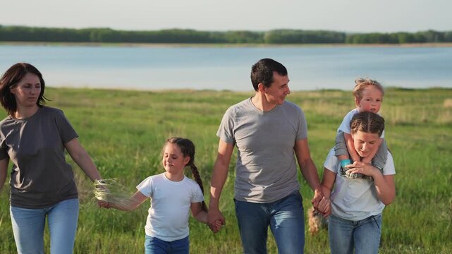 Family walk together by lake shore with father and mother holding daughter and son hand while child sits on parent shoulder in grass field nature setting dog nearby smile and bond happy family unity