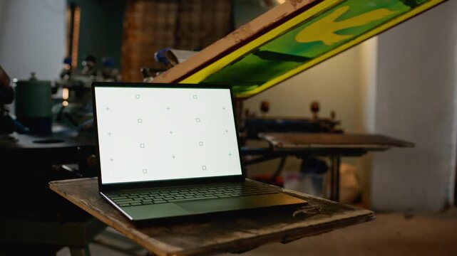No people broll shot of white screen laptop in focus with screen printing machine and frames rotating in workshop background
