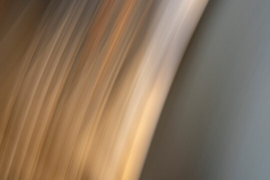 Diagonal streaks of gold and tan light create a motion blur effect against a solid grey background. This abstract visualizes temporal disruption and the fluid volatility of modern systems.