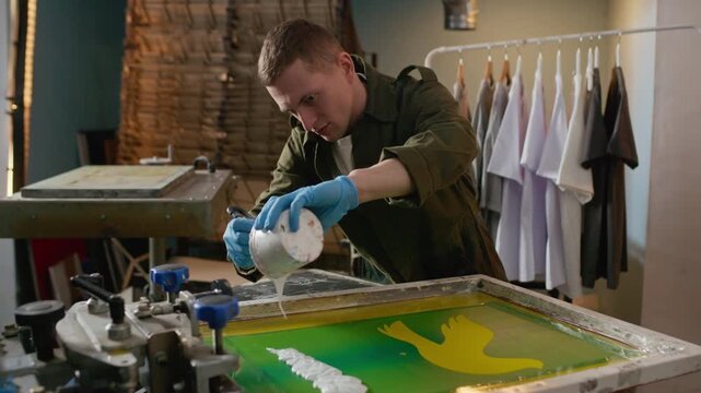 Medium shot of Caucasian male designer in gloves pouring white ink onto screen frame with bird artwork in screen printing workshop