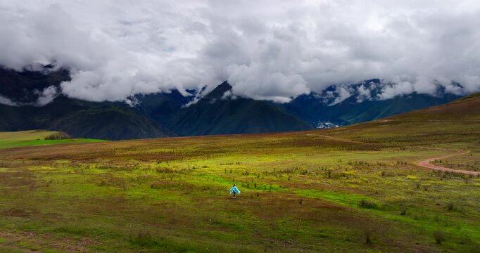 Aerial dolly forward of flamboyant young woman in traditional clothing in the Maras district of the Sacred Valley, Peru. Breathtaking views of the Andean nature and mountains under a cloudy sky