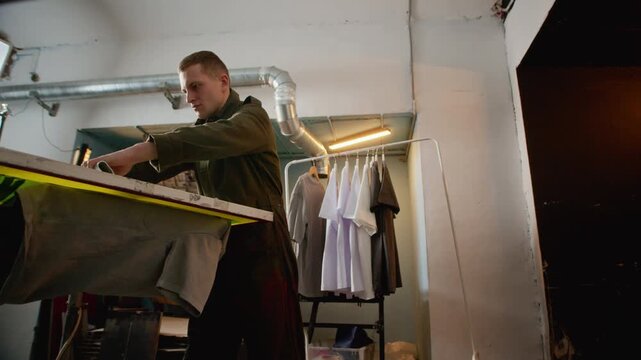 Low angle medium shot of worker spreading ink over screen printing frame using squeegee on press machine in workshop, showing detailed production and apparel printing workflow