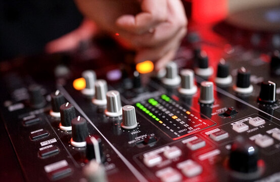 DJ Mixing Music on Professional Controller in Nightclub. Close-up of a DJ's hand adjusting knobs and faders on a professional mixing console. Blurred red bokeh lights in the background