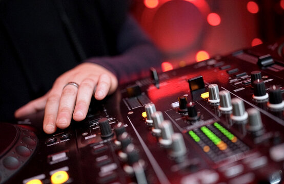 DJ Mixing Music on Professional Controller in Nightclub. Close-up of a DJ's hand adjusting knobs and faders on a professional mixing console. Blurred red bokeh lights in the background
