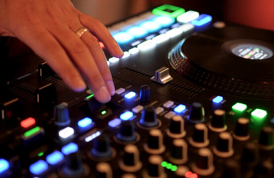 DJ Mixing Music on Professional Controller in Nightclub. Close-up of a DJ's hand adjusting knobs and faders on a professional mixing console. Blurred red bokeh lights in the background