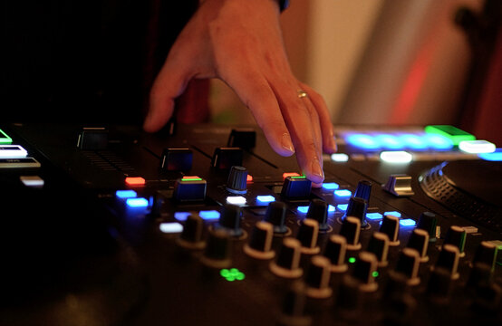 DJ Mixing Music on Professional Controller in Nightclub. Close-up of a DJ's hand adjusting knobs and faders on a professional mixing console. Blurred red bokeh lights in the background