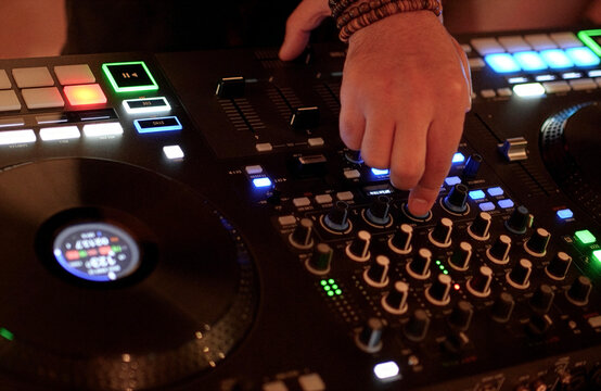 DJ Mixing Music on Professional Controller in Nightclub. Close-up of a DJ's hand adjusting knobs and faders on a professional mixing console. Blurred red bokeh lights in the background