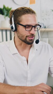 Focused successful young positive businessman sitting at the desk at his workplace during working for laptop, smiling loking at camera at office. Slow morion