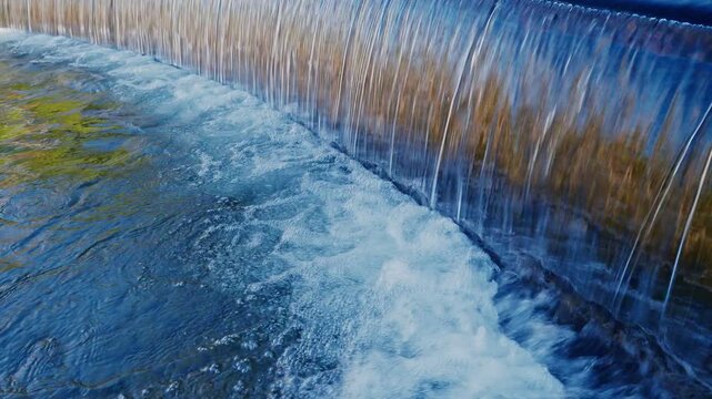 A stunning close-up of water cascading over a curved stone weir in a Japanese garden. The water forms a smooth, glass-like curtain before splashing into the rippling pond below in bright daylight.