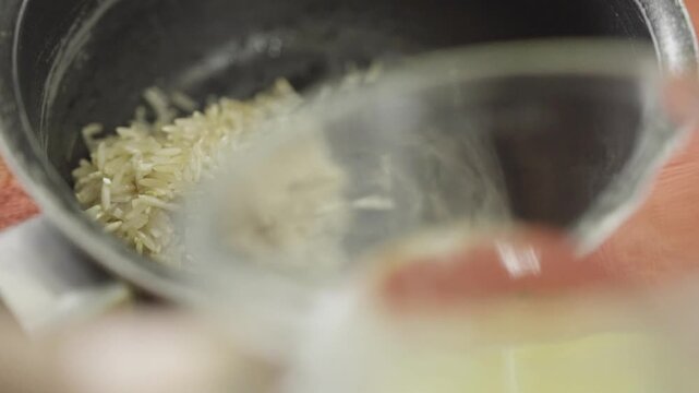 Close up of a hand pouring clear water from a glass into a metal pot containing dry white rice grains, preparing a side dish for albondigas or pollo.