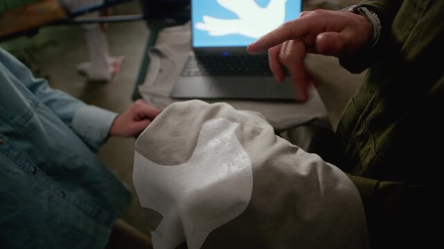 Tracking shot of couple of designers reviewing bird logo on laptop screen while checking matching print on grey t shirt produced with screen printing machine in workshop
