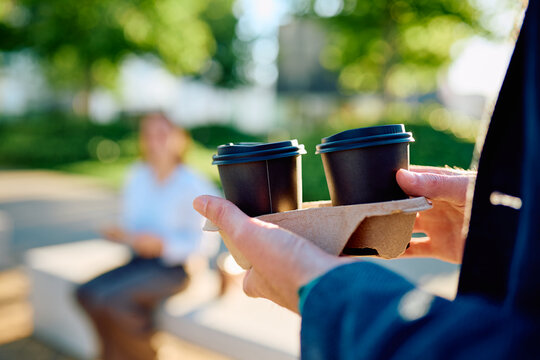 Person carrying two takeaway coffee cups in a holder, delivering drinks to a colleague sitting in a park bench on a sunny day