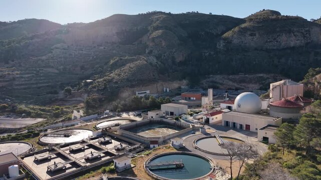 A detailed drone shot showing the EDAR facility, circular clarification tanks, and the prominent spherical biogas storage tank nestled in the mountains.