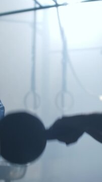 Silhouetted athlete training in misty gym setting with hanging fitness rings, industrial metal frames, and dramatic backlighting creating an intense moody atmosphere