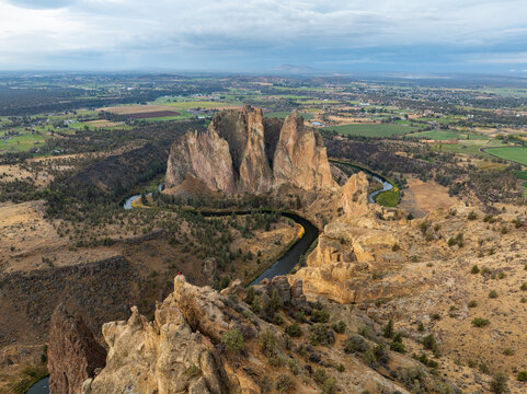 Aerial view of Smith Rock State Park with the winding Crooked River, jagged cliffs, and a person standing on a rocky outcrop under a cloudy sky Crooked River, Oregon, United States.