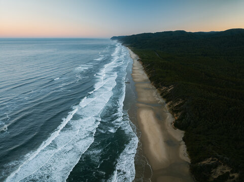 Aerial view of the Oregon Coast Highway winding through dense evergreen forests along a sandy beach with crashing waves at dusk in Florence, Oregon, United States.