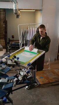 Vertical shot of young Caucasian man using squeegee on screen printing frame in studio workshop, showing creative workflow and hands on production process