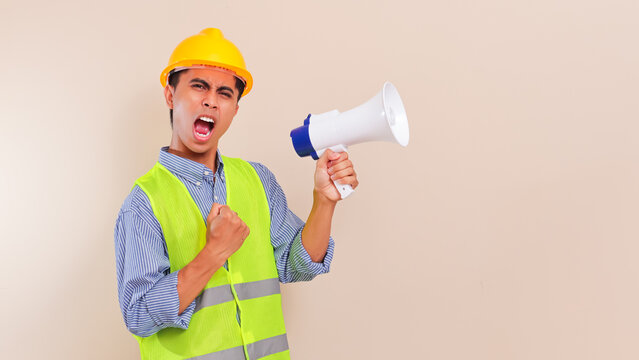 angry young asian construction worker man shouting through megaphone wearing hard hat and safety vest isolated on studio background