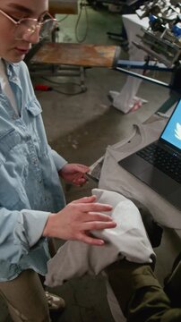 Vertical closeup shot of two workers discussing bird print on grey t shirt created on screen printing machine equipment in creative workshop, checking quality and design