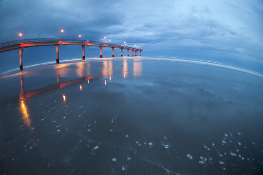 View of a pier stretching into the sea under a dramatic twilight sky, lights reflecting on the wet sand, Brighton Mall, Christchurch, Canterbury Region, New Zealand.