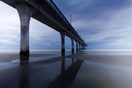 View of a long pier stretching into the calm ocean under a moody sky, its concrete pillars mirrored in the wet sand, Brighton Mall, Christchurch, Canterbury Region, New Zealand.