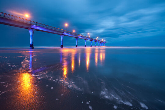 View of a long pier stretching into the sea, illuminated with warm lights that reflect on the wet sand under a dark, moody sky, Christchurch, Canterbury Region, New Zealand.