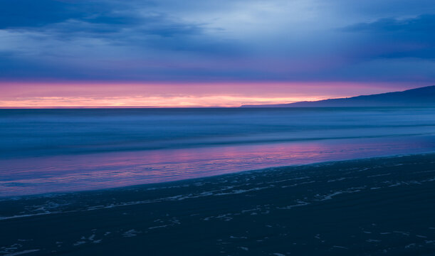 View of the beach reflecting the soft hues of the setting sun, with the landscape silhouette against the horizon, Christchurch, Canterbury Region, New Zealand.
