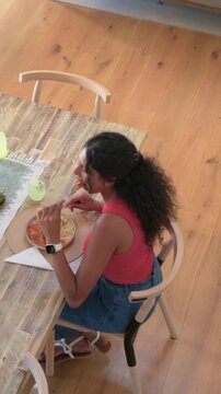 Vertical video: Seated with utensils, woman in coral top and smartwatch cutting food to eat at home